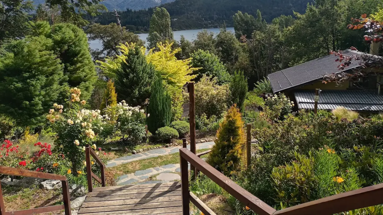 Jardim florido com trilha de madeira e vista para as montanhas e lago, cenário típico da região de Bariloche, Argentina.
