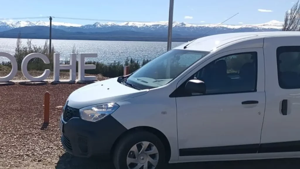 Veículo branco estacionado em frente ao letreiro de Bariloche, com vista para o lago e montanhas nevadas ao fundo.