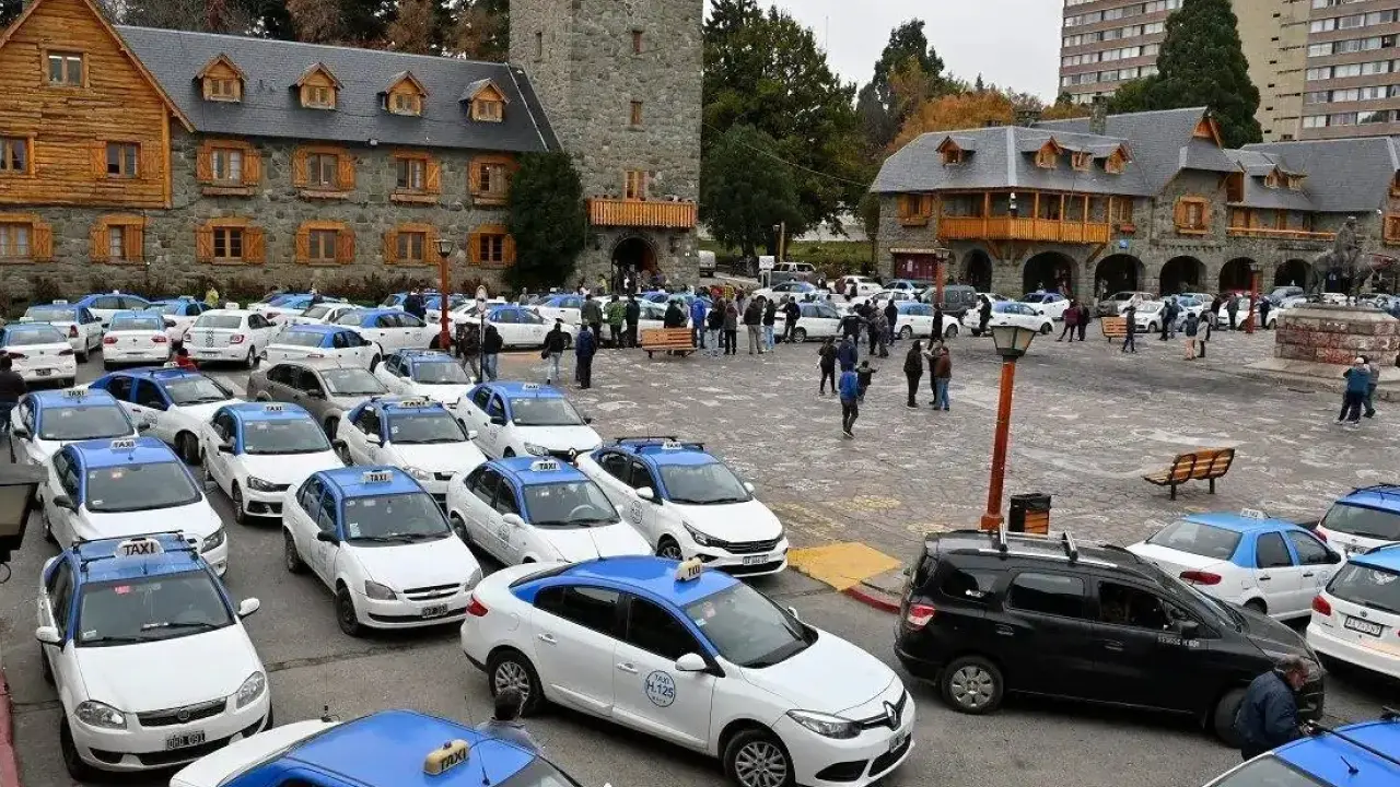 Concentração de táxis brancos com teto azul no Centro Cívico de Bariloche, rodeado por construções em estilo alpino de pedra e madeira.