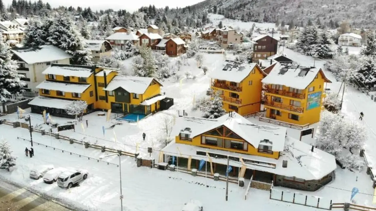 Vista aérea de hospedagens em Bariloche cobertas de neve, com arquitetura alpina e clima típico de inverno patagônico.