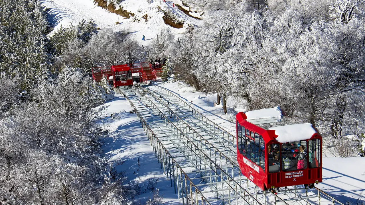 Trilho do Funicular de la Cumbre em Bariloche, cercado por floresta nevada, com vagões vermelhos subindo a montanha.