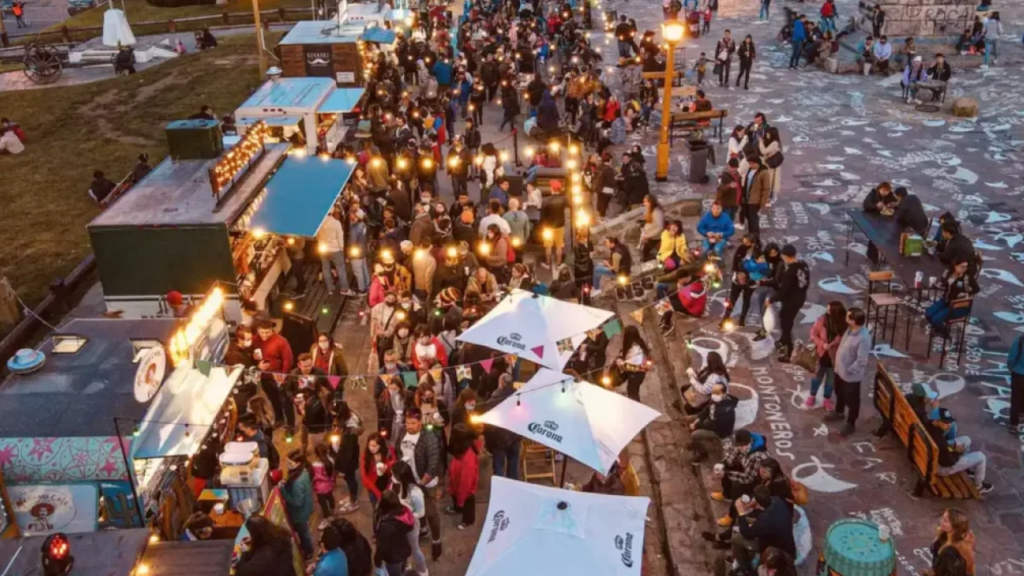 Feira gastronômica ao ar livre com fila de food trucks, mesas comunitárias e luzes decorativas ao entardecer.