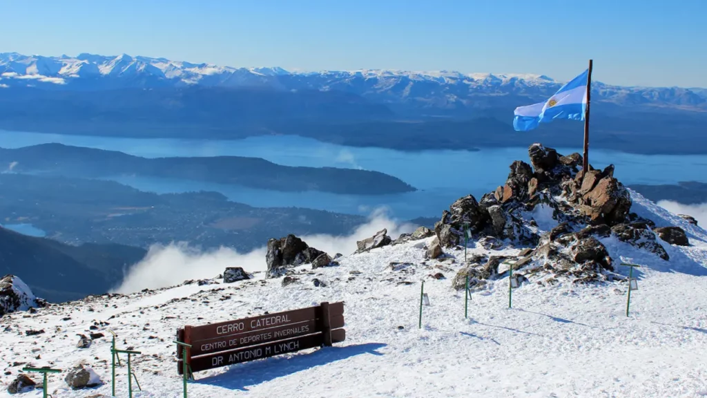 Cume nevado do Cerro Catedral com bandeira da Argentina e lagos ao fundo.