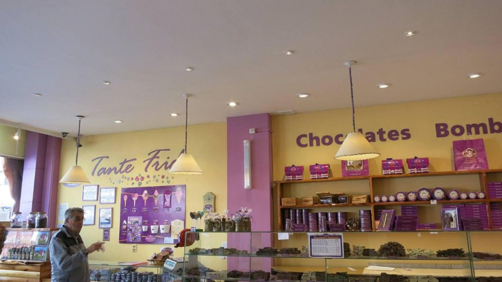 Interior de chocolateria com vitrines de doces, paredes amarelo-lilás e cliente ao lado esquerdo.