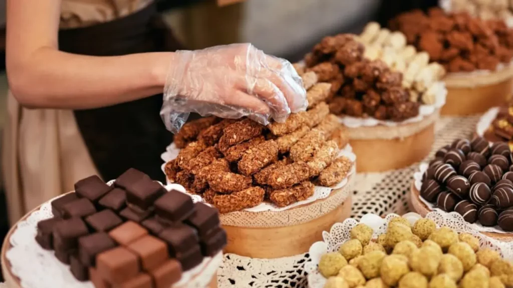 Bandejas com variedades de chocolates artesanais e alfajores, com mão enluvada organizando os doces em uma loja típica de Bariloche.
