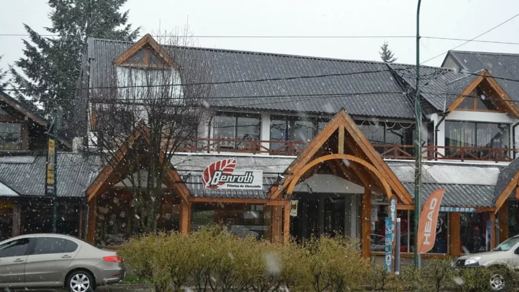 Fachada da chocolateria Benroth em Bariloche durante uma leve nevasca, com arquitetura de madeira alpina e letreiro visível.