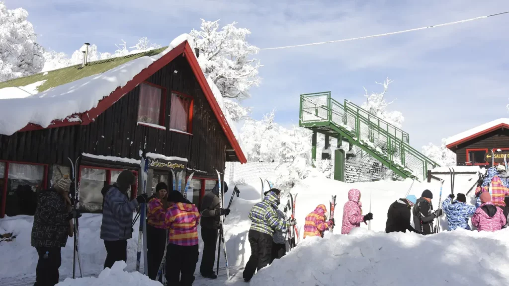 Visitantes equipados para atividades na neve em Piedras Blancas, Bariloche, com chalés cobertos de neve e escadas de acesso verde.