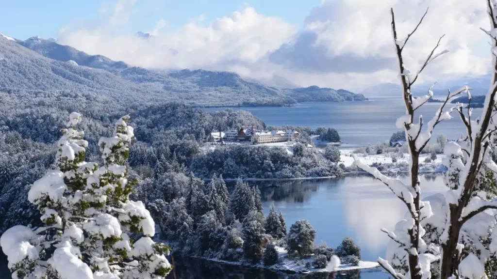 Vista panorámica invernal de Bariloche con bosques nevados, un lago cristalino y el Hotel Llao Llao en el centro del paisaje.