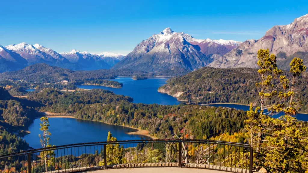 Vista desde un mirador en el Circuito Chico de Bariloche, con lagos azules, montañas andinas nevadas y bosque en tonos verdes y otoñales.