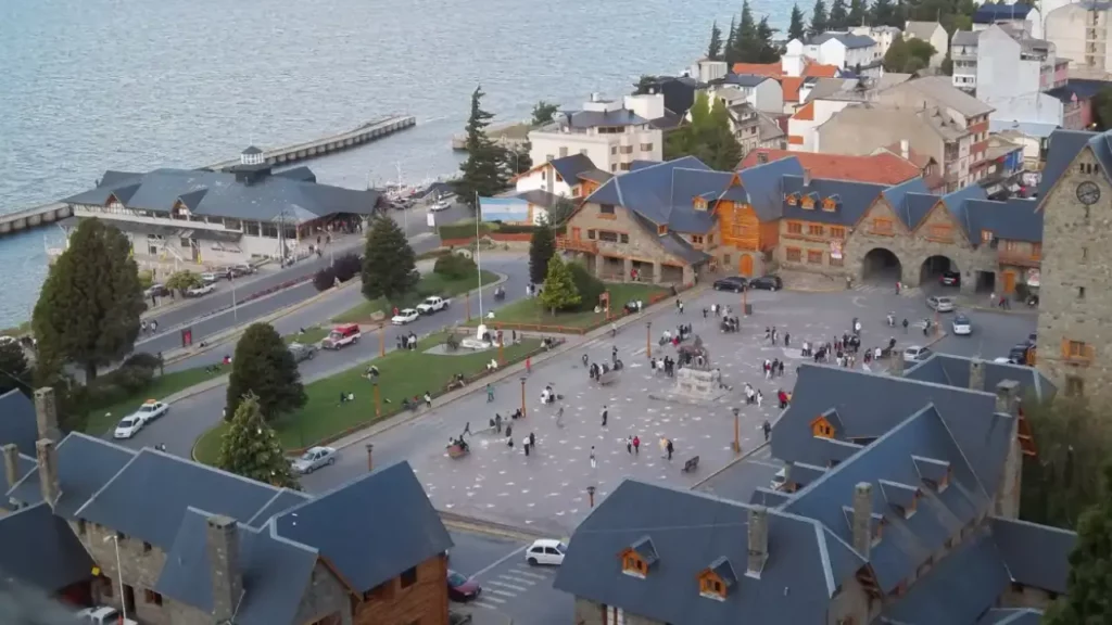Vista aérea da praça principal de Bariloche, com construções em estilo alpino, turistas caminhando e o lago Nahuel Huapi ao fundo.