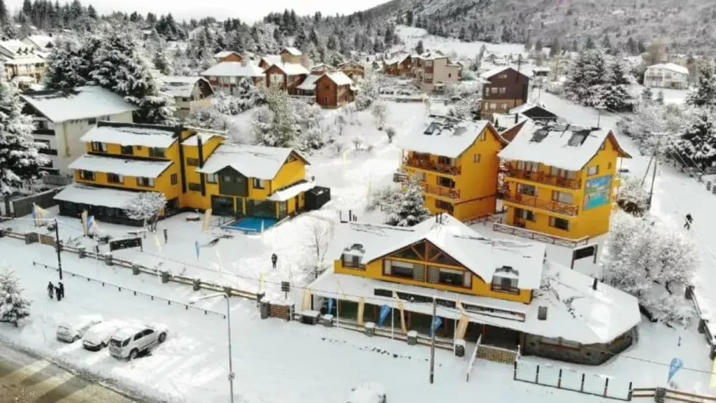 Vista aérea de hospedagens em Bariloche cobertas de neve, com arquitetura alpina e clima típico de inverno patagônico.