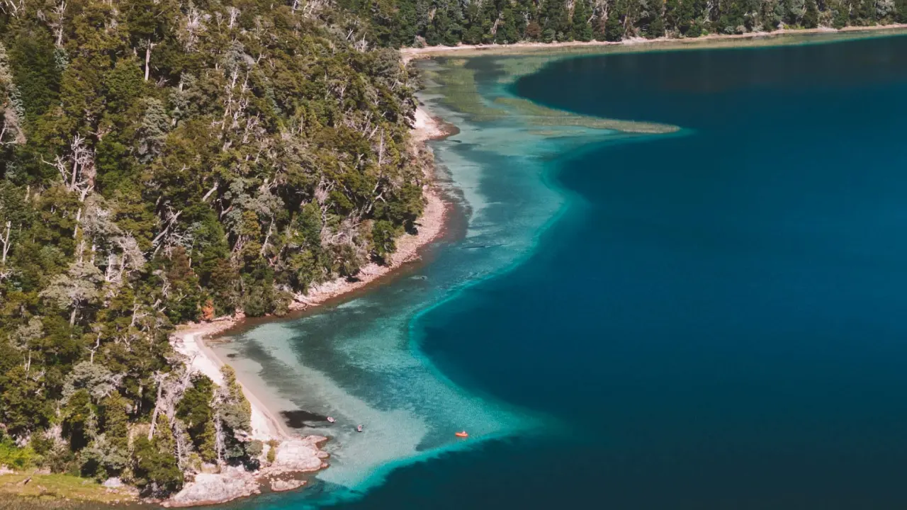 Vista aérea de um lago em Bariloche, mostrando a transição da água de um azul-turquesa cristalino para um azul profundo, com uma floresta densa na encosta.