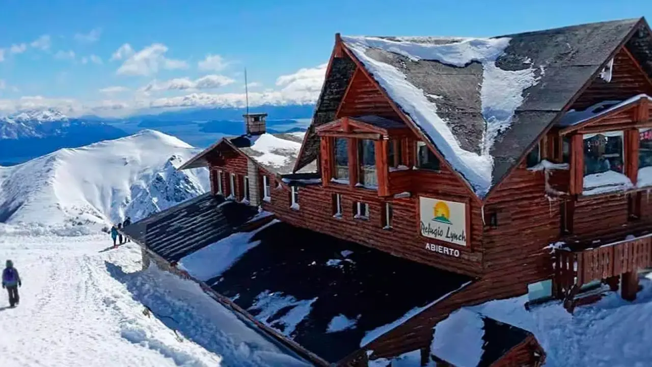 Refúgio de madeira com telhado nevado no Cerro Catedral, Bariloche, com vista para montanhas cobertas de neve e céu azul intenso.