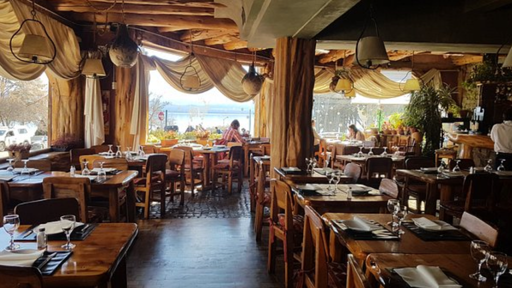 Interior de restaurante com decoração rústica em madeira, mesas bem postas e vista para o lago ao fundo, transmitindo aconchego e elegância.