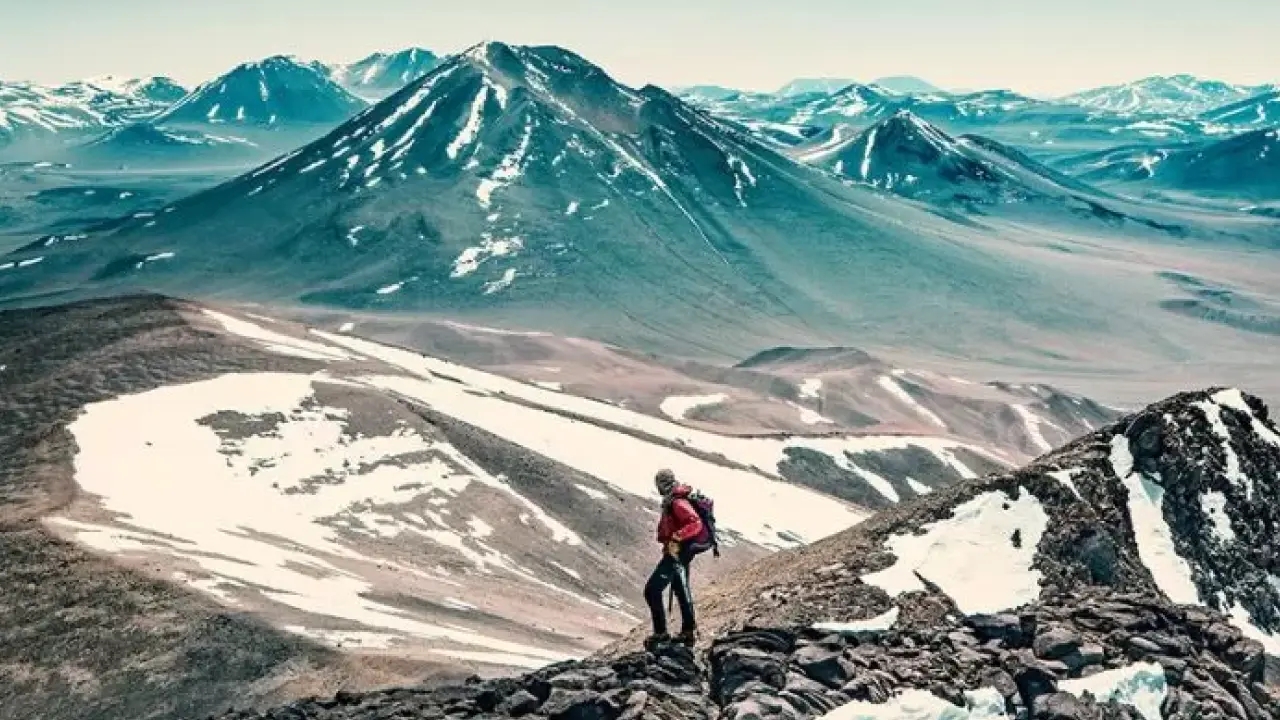 Trekker com mochila no cume de uma montanha rochosa, observando uma vasta paisagem de picos com neve ao fundo.