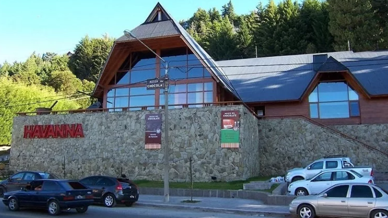 Vista frontal da Fábrica de Chocolate Havanna em Bariloche com carros estacionados na rua e floresta ao fundo.