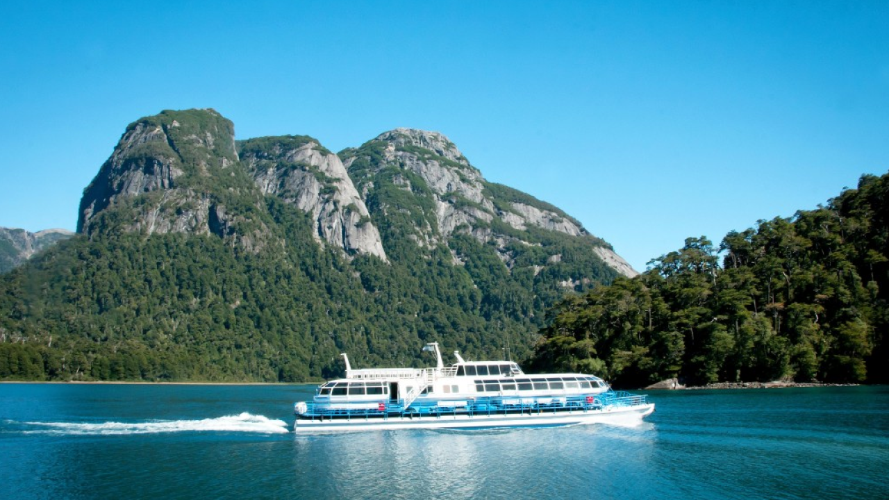 Barco de passeio navegando pelo Lago Todos los Santos, com montanhas cobertas por vegetação ao fundo, em um dia ensolarado na Patagônia chilena.