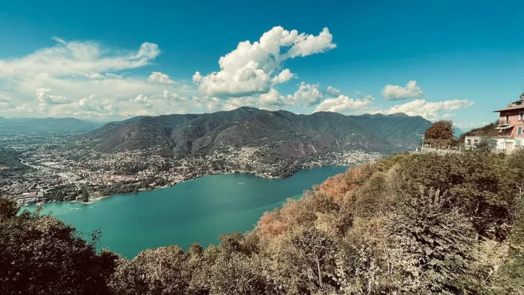 Vista panorâmica do Lago de Como cercado por montanhas e cidadezinhas italianas em um dia claro de céu azul e nuvens leves.