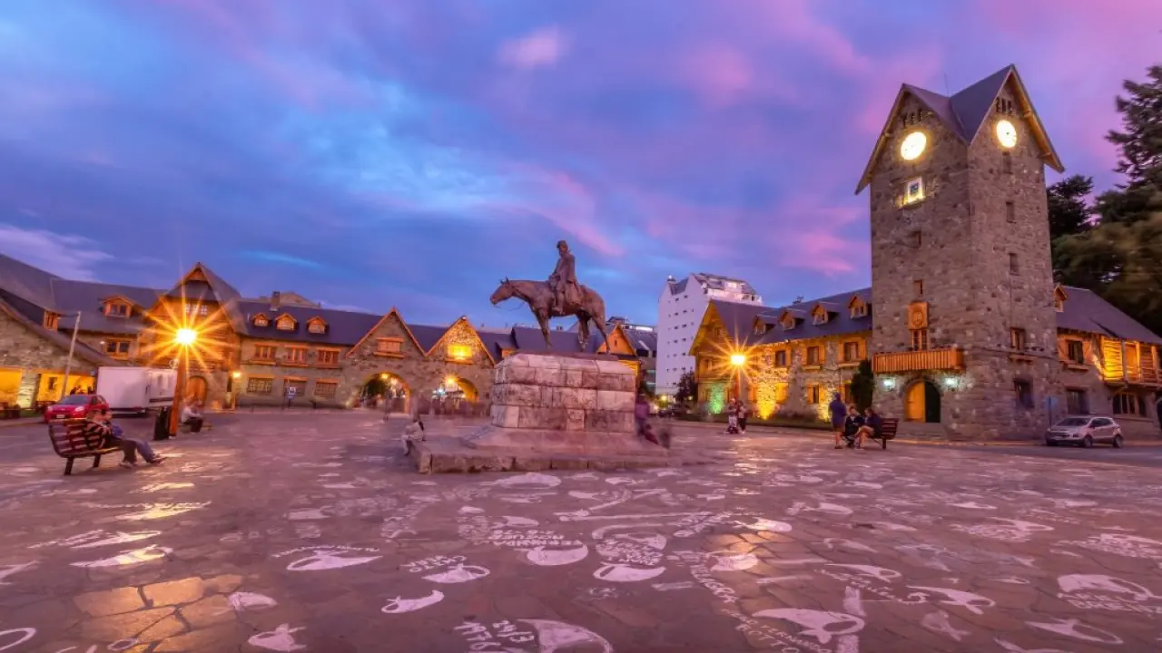 Praça principal ao entardecer com um céu em tons de roxo e rosa. Ao centro, uma estátua equestre sobre um pedestal de pedra. Ao fundo, construções de pedra e madeira com relógios nas torres, em estilo alpino. No chão, há desenhos de mãos e frases pintadas.