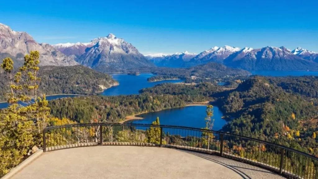 Mirante com vista panorâmica para lagos azuis cercados por florestas e montanhas cobertas de neve. O céu está limpo e azul, sugerindo um dia ensolarado. A cena transmite tranquilidade e beleza natural exuberante.