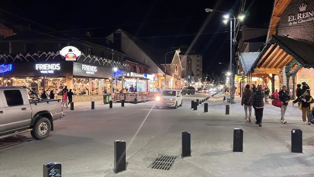 Rua movimentada à noite em uma cidade com iluminação artificial. Lojas e restaurantes, como o "FRIENDS", estão iluminados e cheios de pessoas. Carros circulam pela via, e pedestres caminham nas calçadas. À direita, há um prédio com o nome "El Reino de los Chocolates".