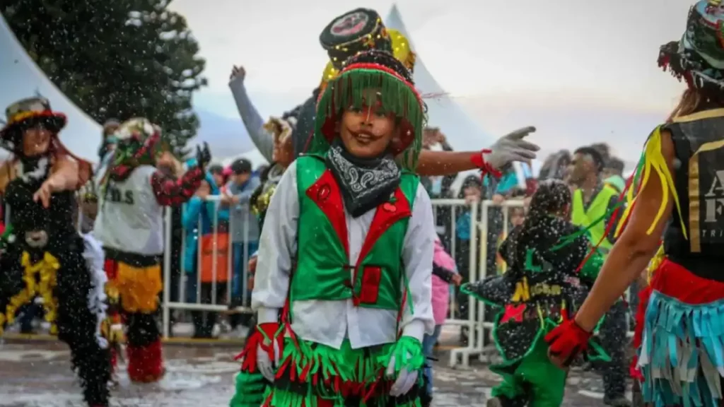 Criança com fantasia colorida participa de desfile folclórico durante o Carnaval de Inverno em Bariloche, com neve artificial e público ao fundo.