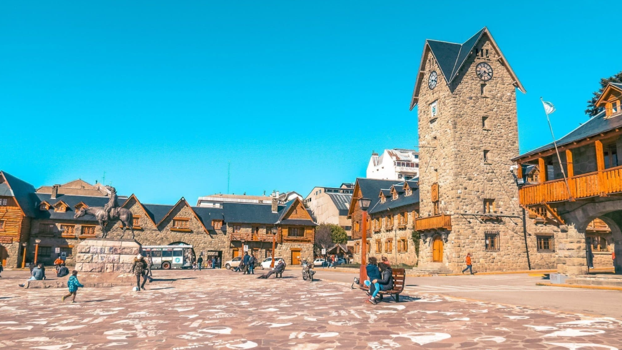 Praça do Centro Cívico de Bariloche com turistas, prédios de pedra e a famosa torre do relógio em um dia claro.