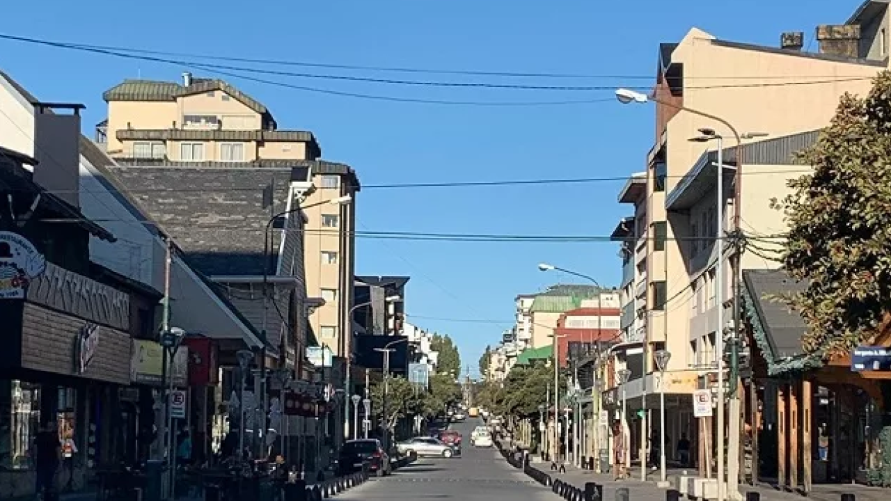 Rua principal de Bariloche em um dia ensolarado, com prédios baixos, comércios e trânsito leve.