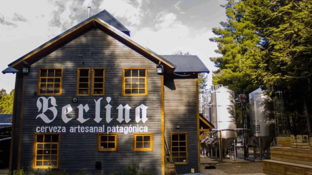 Fachada rústica da cervejaria Berlina, com a inscrição 'cerveza artesanal patagónica', cercada por pinheiros e tanques de produção ao fundo."