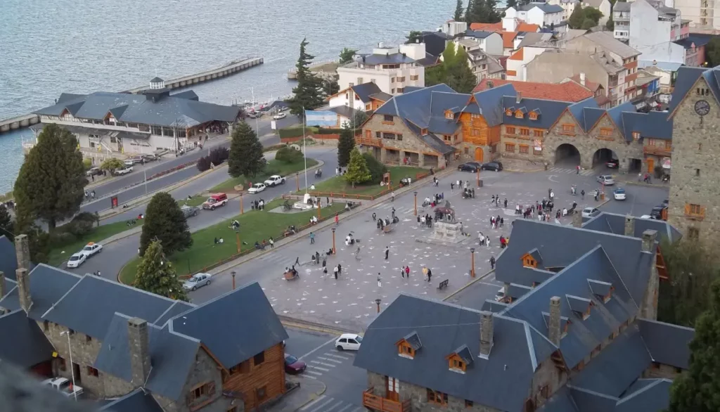 Vista aérea da praça principal de Bariloche com o lago Nahuel Huapi ao fundo, cercada por edifícios com arquitetura alpina.