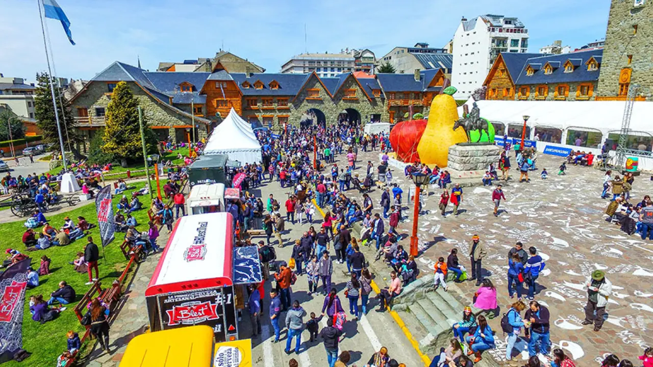 Multidão caminha pela praça cívica de Bariloche durante evento ao ar livre com food trucks e esculturas coloridas gigantes.