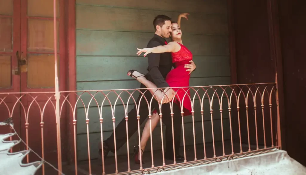 Um casal dançando tango em uma varanda com grades de ferro, com o homem vestindo terno preto e a mulher em um vestido vermelho. A dança elegante e apaixonada é parte do Festival Internacional de Tango e Patagônia em Bariloche.