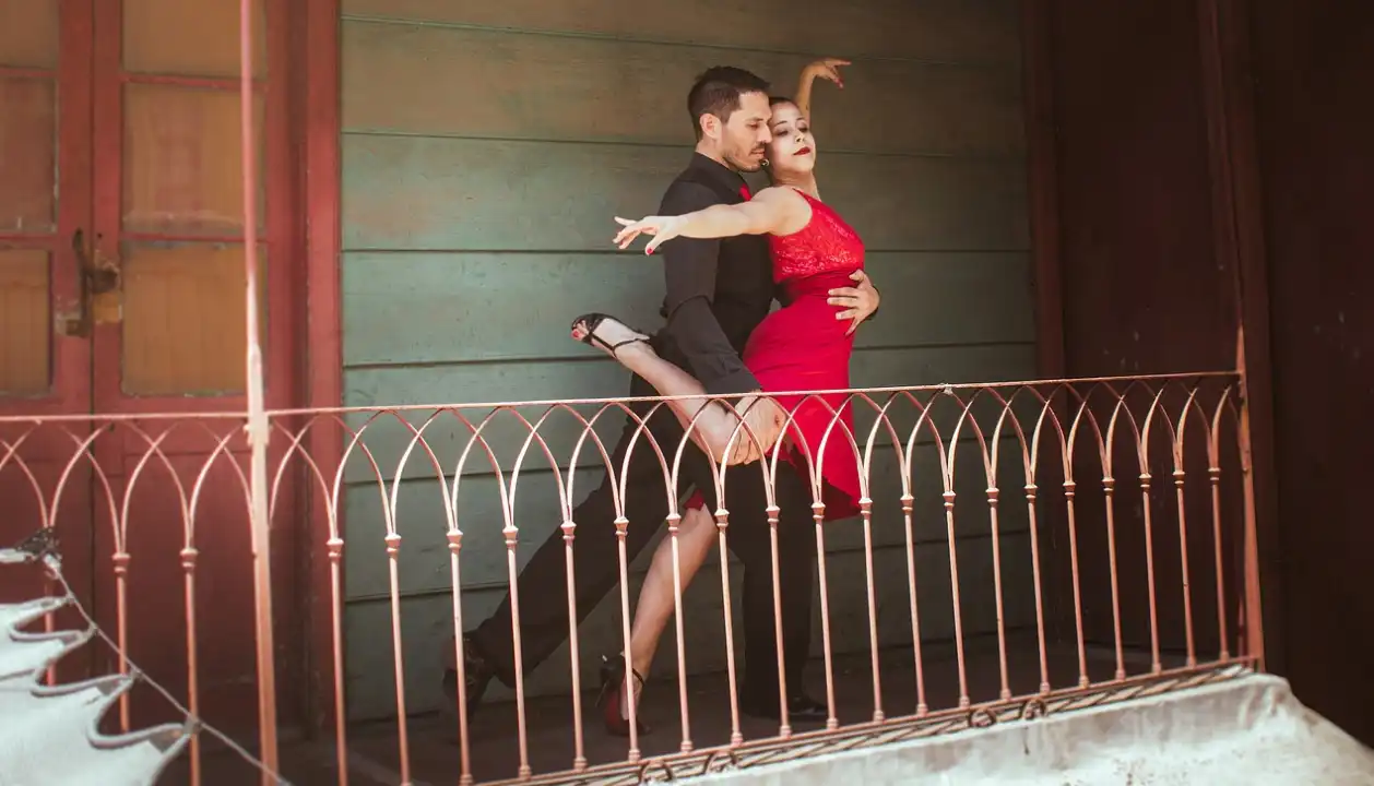 Um casal dançando tango em uma varanda com grades de ferro, com o homem vestindo terno preto e a mulher em um vestido vermelho. A dança elegante e apaixonada é parte do Festival Internacional de Tango e Patagônia em Bariloche.