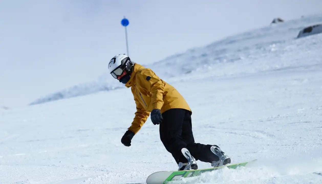 Uma pessoa praticando snowboard em uma montanha coberta de neve, vestindo uma jaqueta amarela e capacete branco. O céu está claro e a paisagem nevada cria um ambiente perfeito para esportes de inverno.