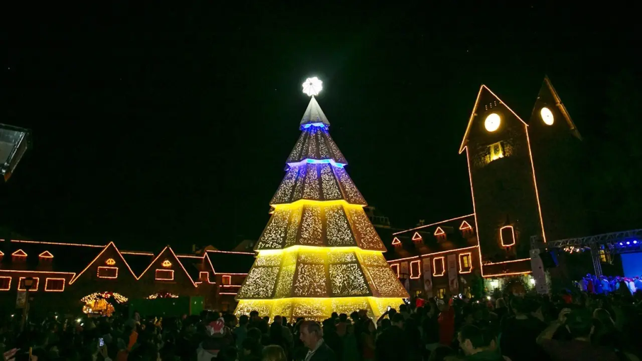 Árvore de Natal iluminada na praça principal de Bariloche durante a celebração natalina, cercada por turistas à noite.