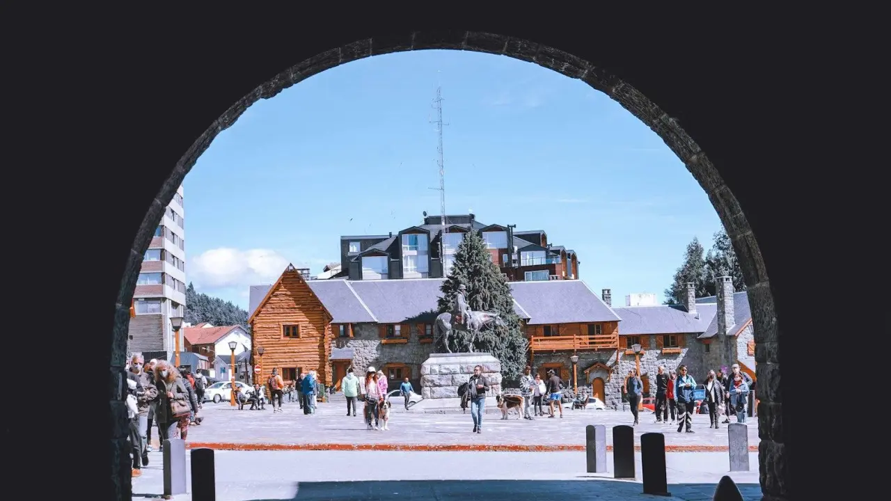 Praça principal de Bariloche vista através de um arco, com pessoas passeando e edifícios históricos ao fundo.