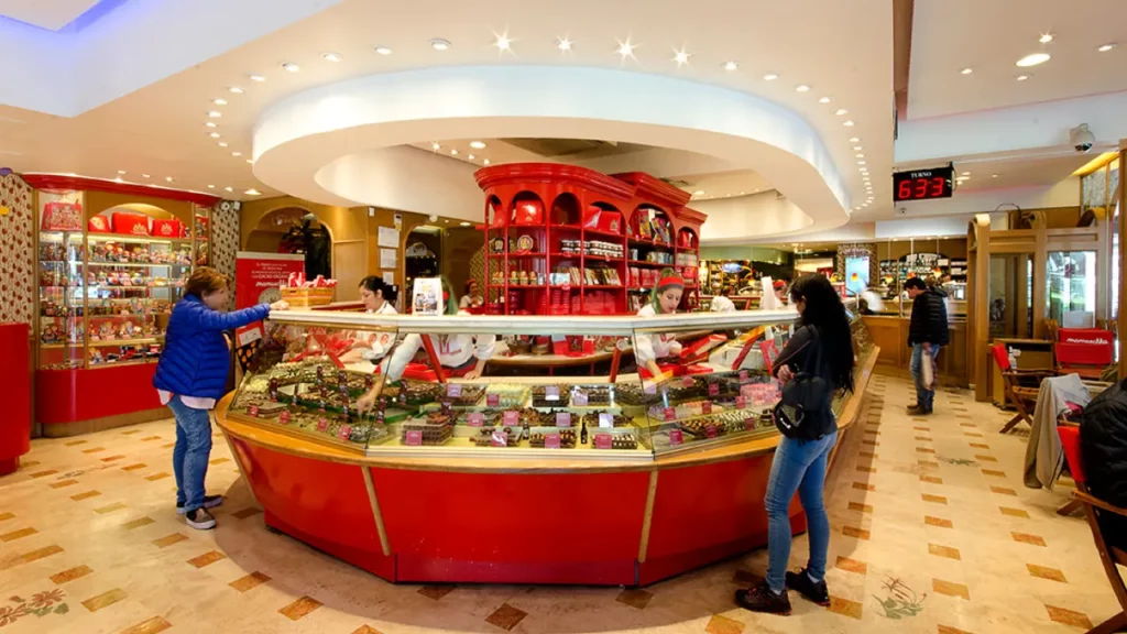 Interior da loja Mamuschka com balcão circular de chocolates, decoração em vermelho, atendentes uniformizadas e clientes sendo servidos.