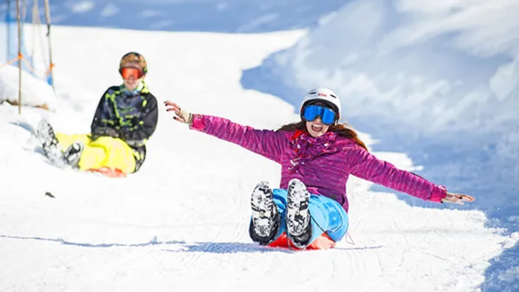 Jovens se divertindo ao descer na neve com trenós no parque Piedras Blancas, em Bariloche, usando roupas de inverno e capacete.