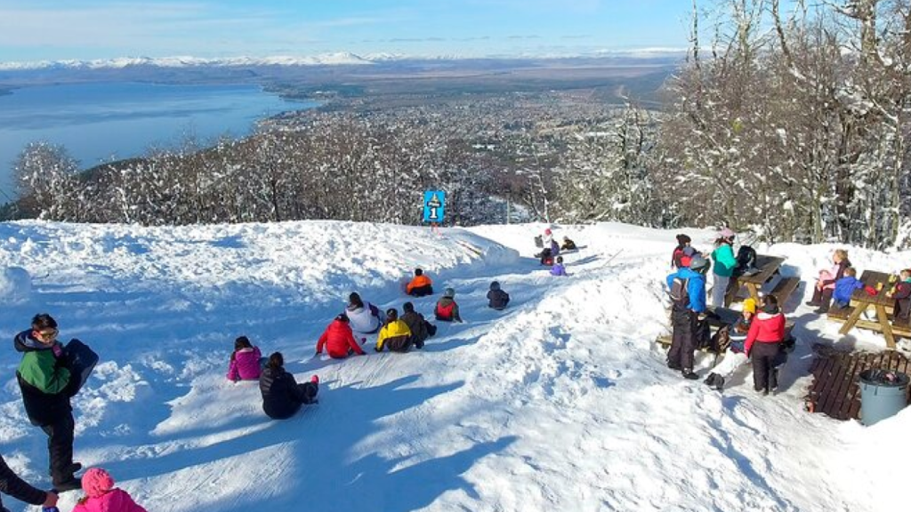 Grupo de visitantes descendo em tobogãs de neve no parque Piedras Blancas, com vista panorâmica do lago Nahuel Huapi e da cidade de Bariloche ao fundo.