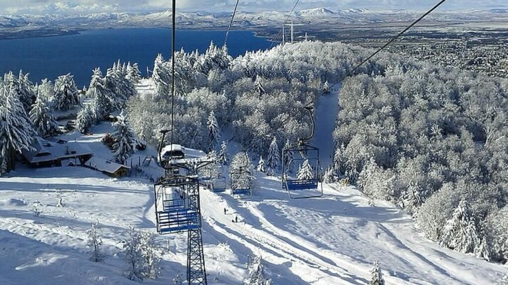 Cadeira de teleférico subindo entre árvores cobertas de neve no parque Piedras Blancas, com vista do lago e das montanhas de Bariloche ao fundo.