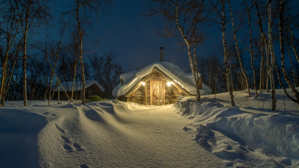 Cabana de madeira coberta de neve com luzes acesas na entrada, cercada por árvores sob o céu noturno.