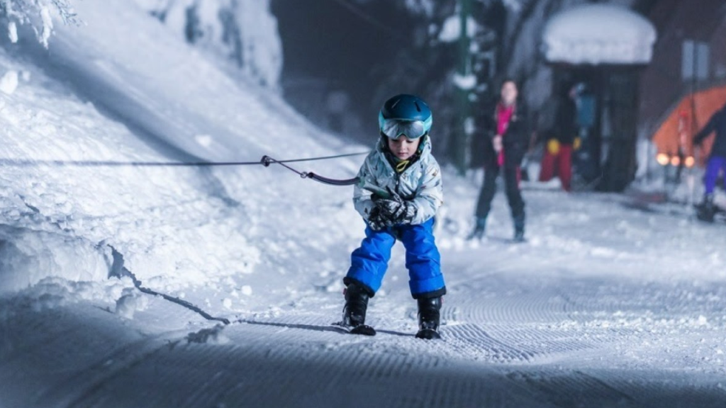 Criança com roupa de neve sendo puxada por corda em pista de esqui à noite, usando capacete e óculos de proteção.