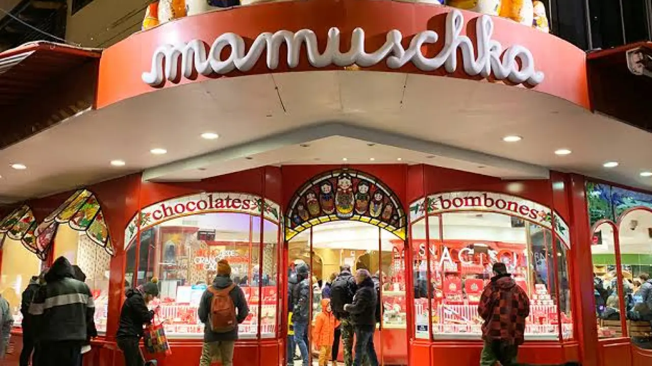 Fachada iluminada da loja Mamuska em Bariloche, com várias bonecas russas decorando o topo e uma vitrine cheia de chocolates artesanais e bombons. Pessoas entram e saem da loja enquanto apreciam as delícias oferecidas.