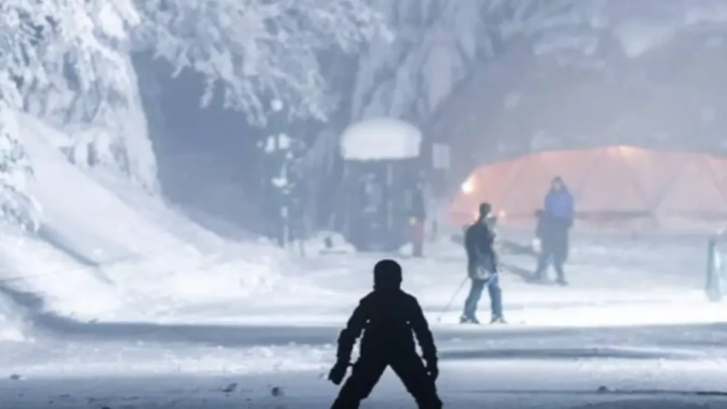 Pessoa esquiando à noite em uma pista iluminada, cercada por árvores cobertas de neve, com algumas pessoas ao fundo perto de uma estrutura iluminada em um ambiente de inverno.