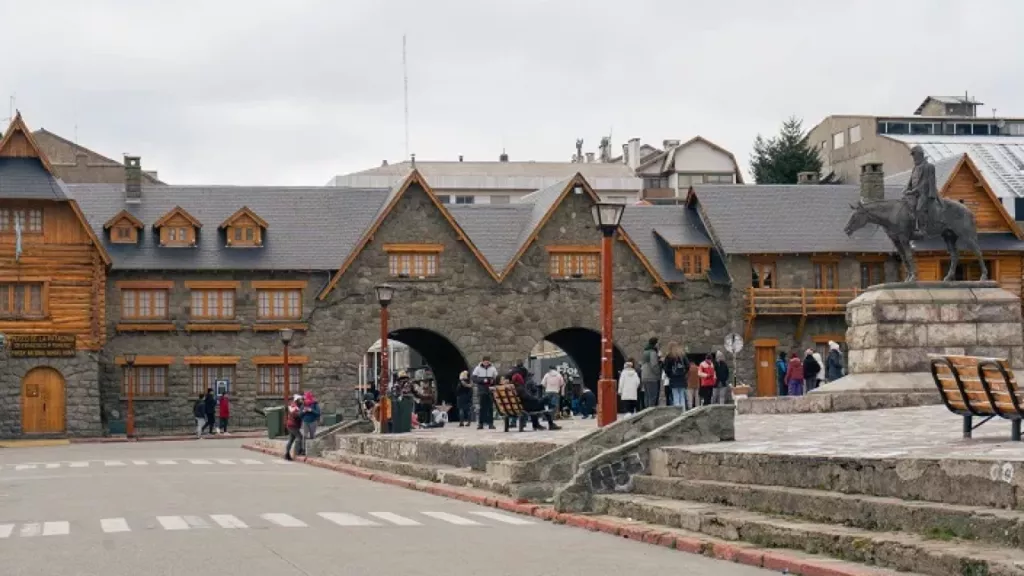 Escadaria e estátua do Centro Cívico de Bariloche com céu nublado e visitantes reunidos.