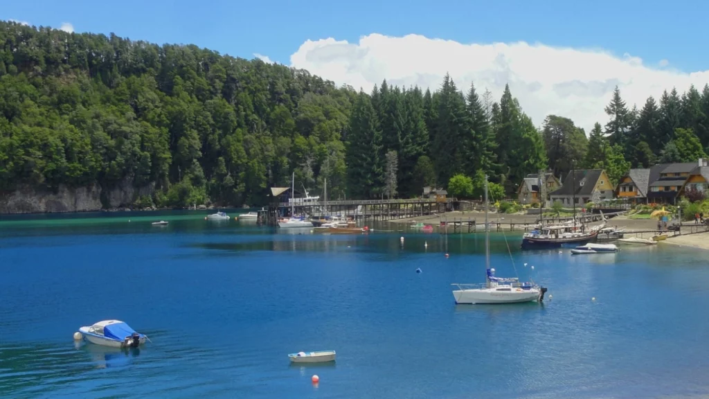 Uma praia calma em Bariloche com barcos ancorados na água azul-turquesa, cercada por árvores e chalés. Um ambiente perfeito para atividades ao ar livre durante a primavera.