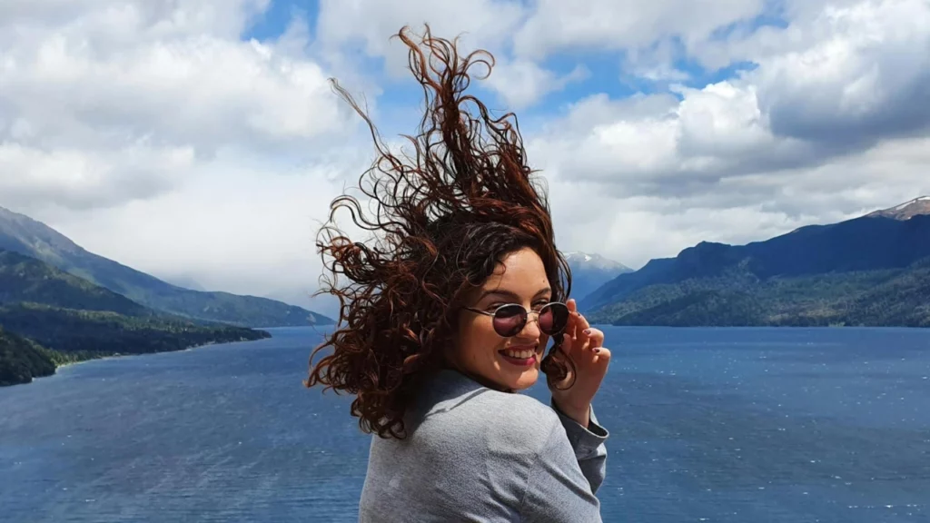 Uma jovem sorridente com cabelo ao vento, usando óculos de sol, em um mirante de Bariloche. Ao fundo, montanhas verdes e o vasto lago azul, capturando o espírito livre e as paisagens deslumbrantes da primavera.