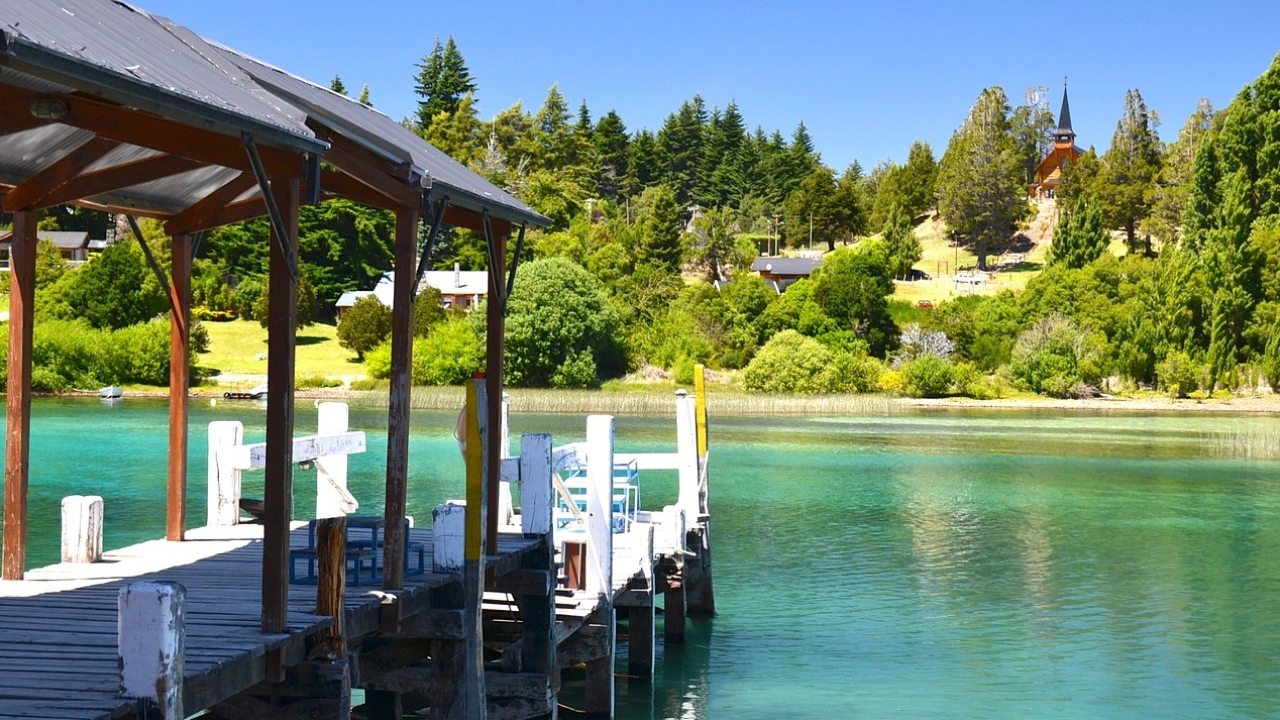 Um píer de madeira em uma água cristalina azul-turquesa em Bariloche, rodeado por árvores verdes e com uma igreja ao fundo. O cenário tranquilo destaca a beleza natural da primavera na região.