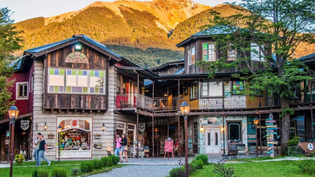 Rua charmosa em Villa La Angostura com arquitetura de madeira e pedra, destacando lojas de artesanato local e turistas caminhando pelas calçadas. Ao fundo, uma montanha iluminada pela luz do sol.