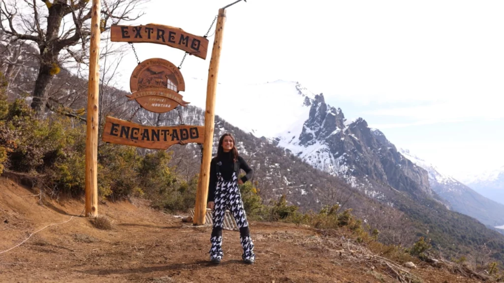 Mulher posando em frente a uma placa de madeira que diz 'Extremo Encantado', com uma paisagem montanhosa coberta de neve ao fundo e o céu parcialmente nublado.